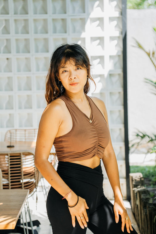 A woman posing in a brown ruched halter crop top and black leggings in an outdoor patio setting with a white glass block wall background.