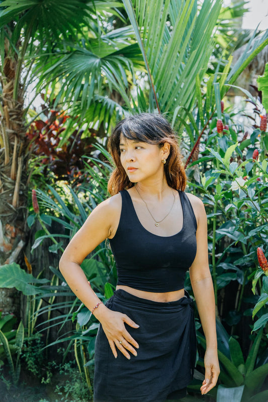 A woman wearing a black ribbed tank top and matching black skirt stands in a lush tropical garden setting.
