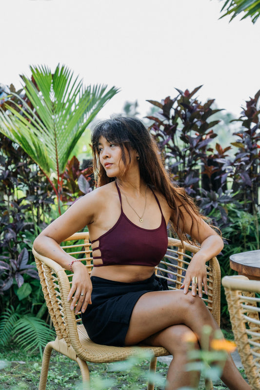 A woman sitting on a woven outdoor chair wearing a burgundy halter crop top and black shorts, posed in a lush garden setting.