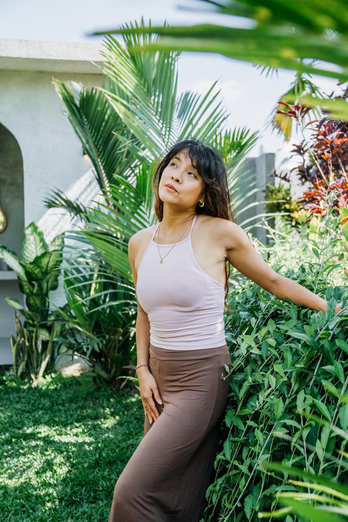 A woman posing outdoors in a garden setting, wearing a light pink ribbed tank top and brown wide-leg pants.