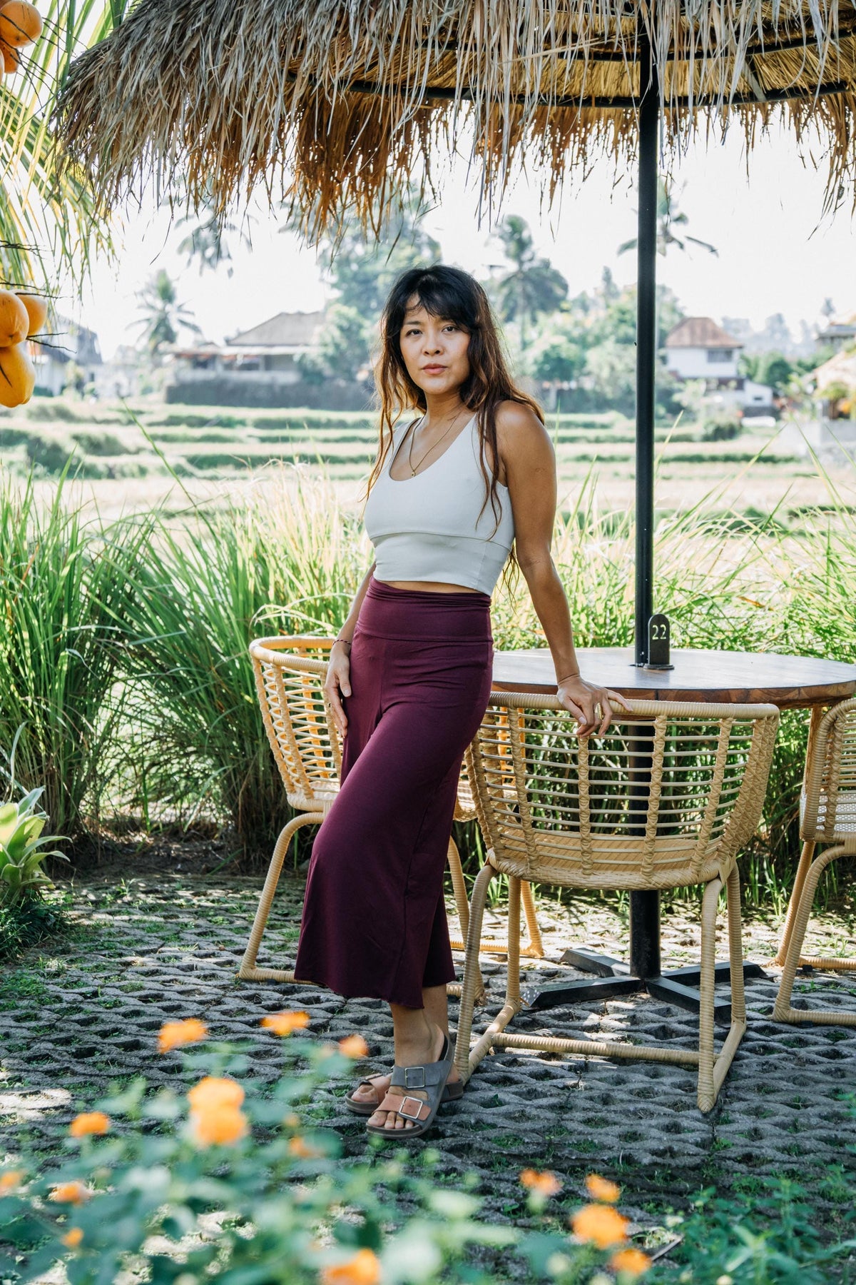 A woman stands outdoors at a table under a thatched umbrella, wearing a light beige sleeveless crop top and a long burgundy maxi skirt. The setting features woven chairs, a stone floor, and a lush green rice field in the background.