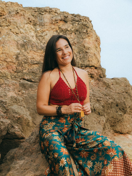 A woman sitting outdoors on large tan rocks, wearing a red crochet halter top and a teal floral maxi skirt, with long beaded necklaces and bracelets.