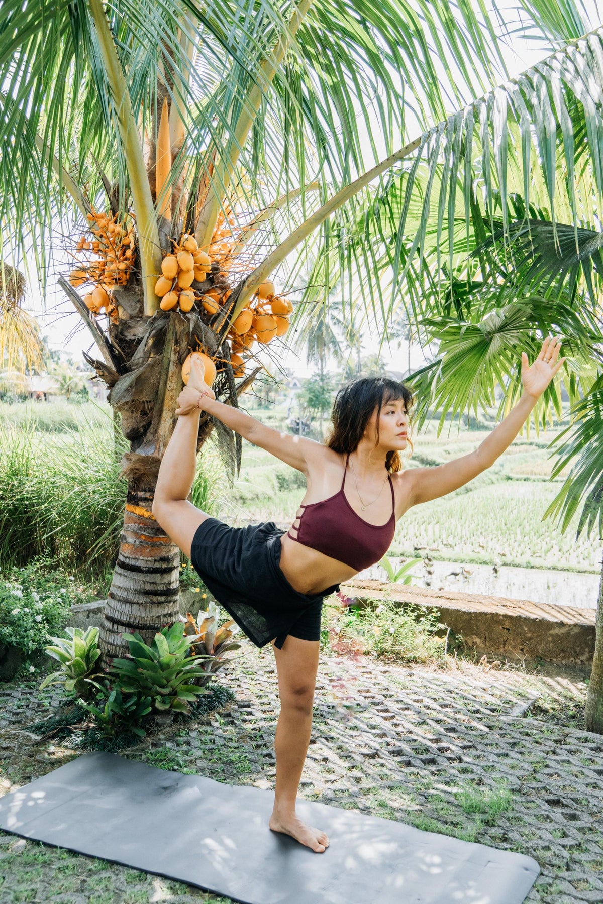 A woman practices yoga outdoors in a tropical garden, balancing in a side stretch pose on a dark yoga mat next to a palm tree with coconuts. She is wearing a maroon sports bra and black shorts.