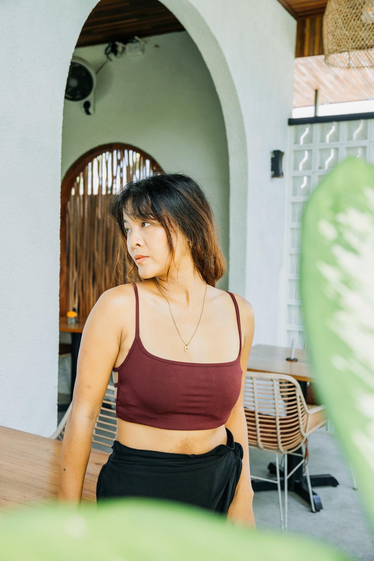 A woman wearing a burgundy ribbed crop top and black bottoms, standing in a modern outdoor patio setting with arches and wooden accents.