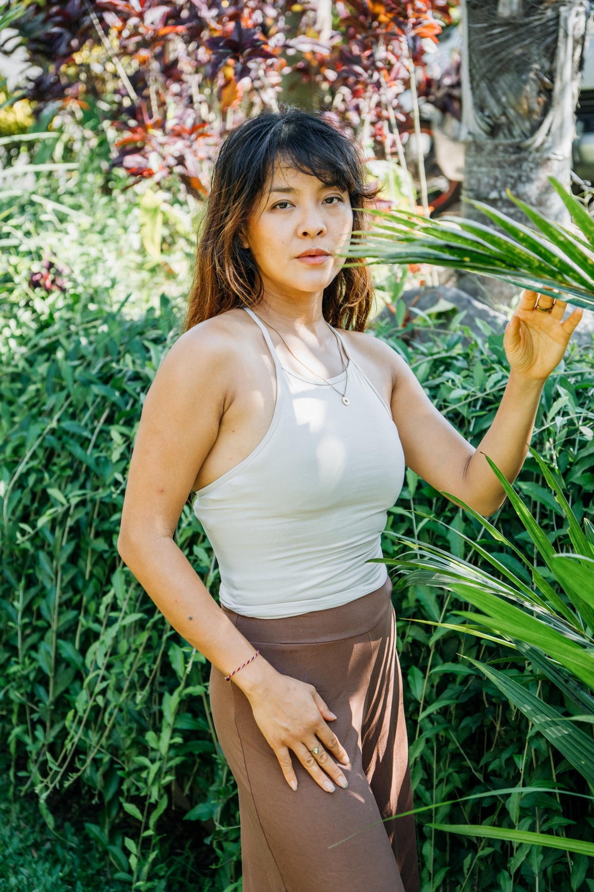 A woman posing outdoors in a garden, wearing a light beige ribbed tank top and brown pants, surrounded by lush green foliage.