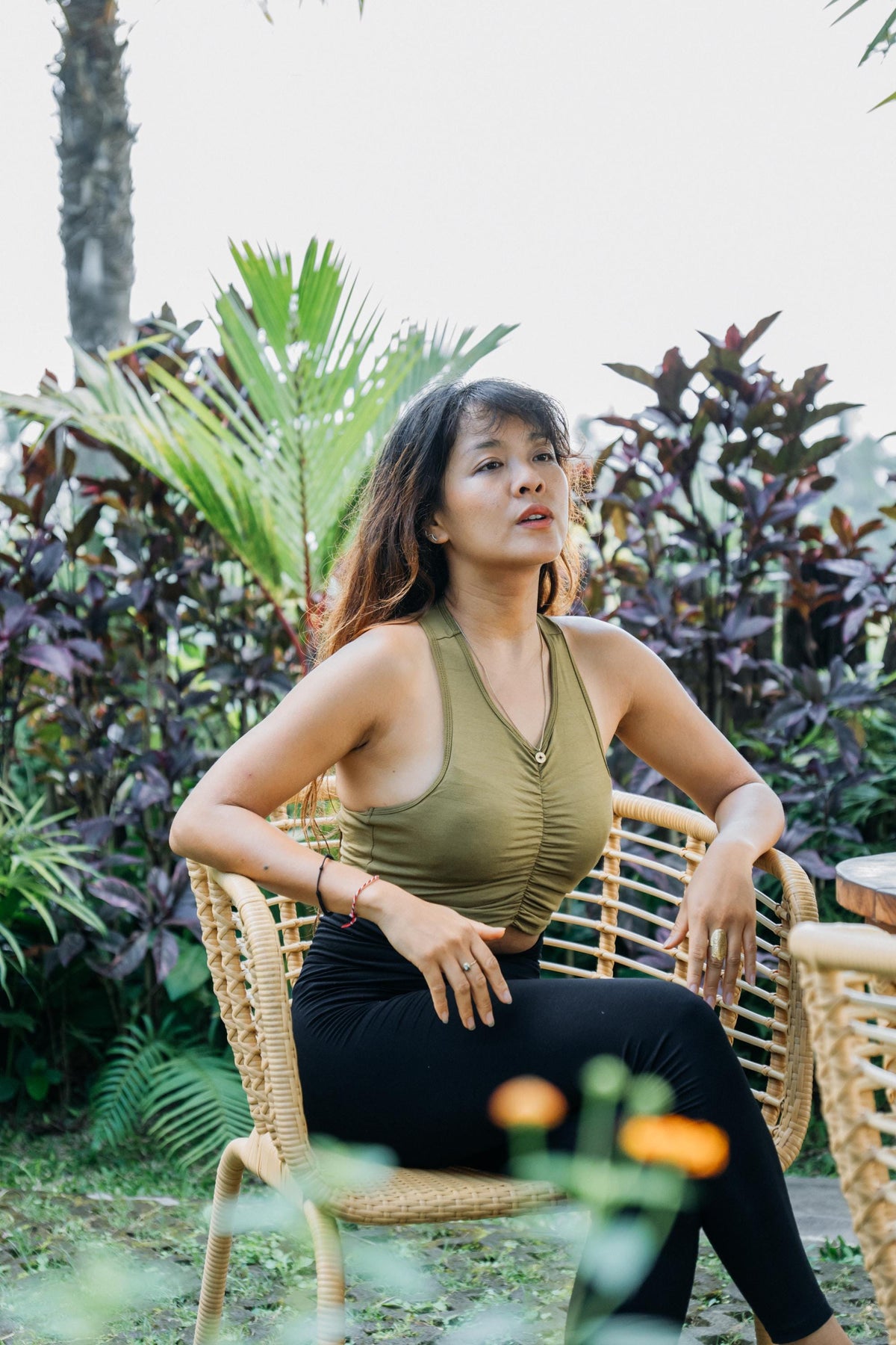 A woman sitting in a wicker chair outdoors, wearing a ruched olive green tank top and black leggings, with tropical plants in the background.