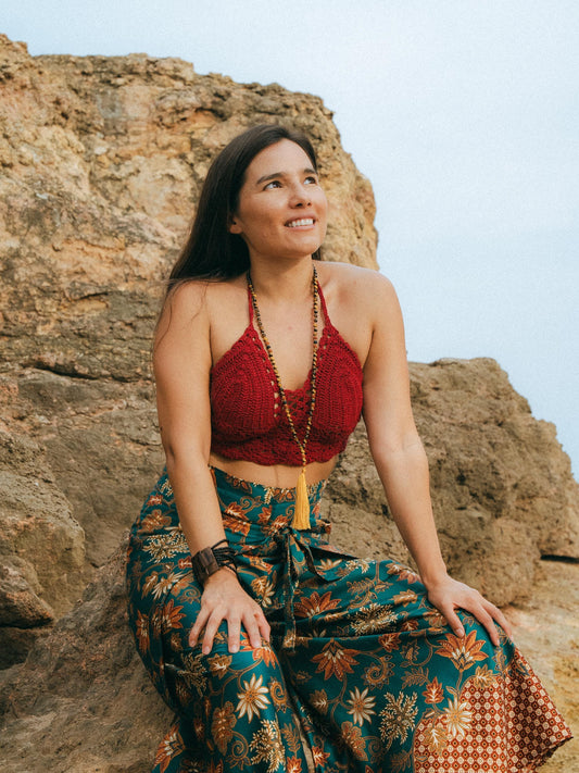A woman sitting on a rocky cliffside wearing a red crochet halter crop top and a teal floral maxi skirt with gold patterns, accessorized with a long beaded tassel necklace.