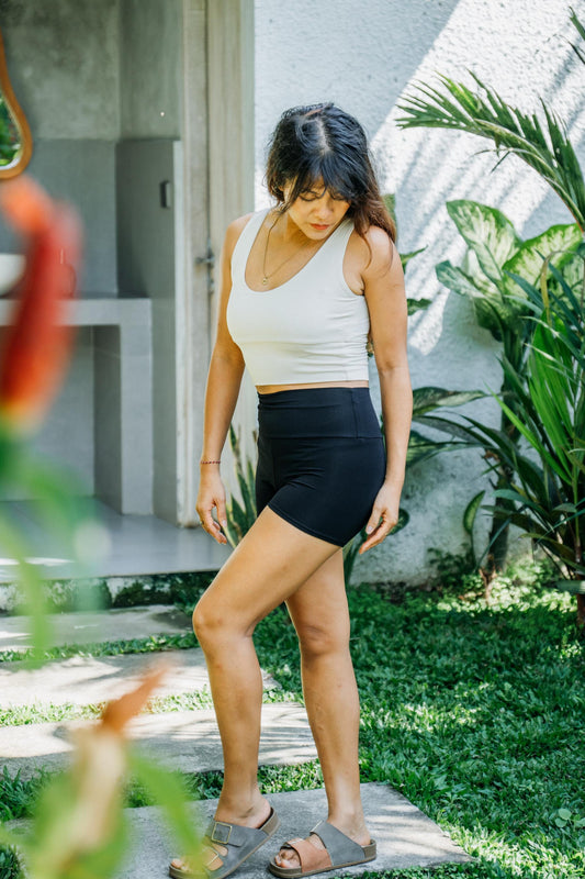 A woman wearing a white ribbed tank top and black high-waisted shorts stands outdoors on a stone path with green plants and a stucco wall in the background.