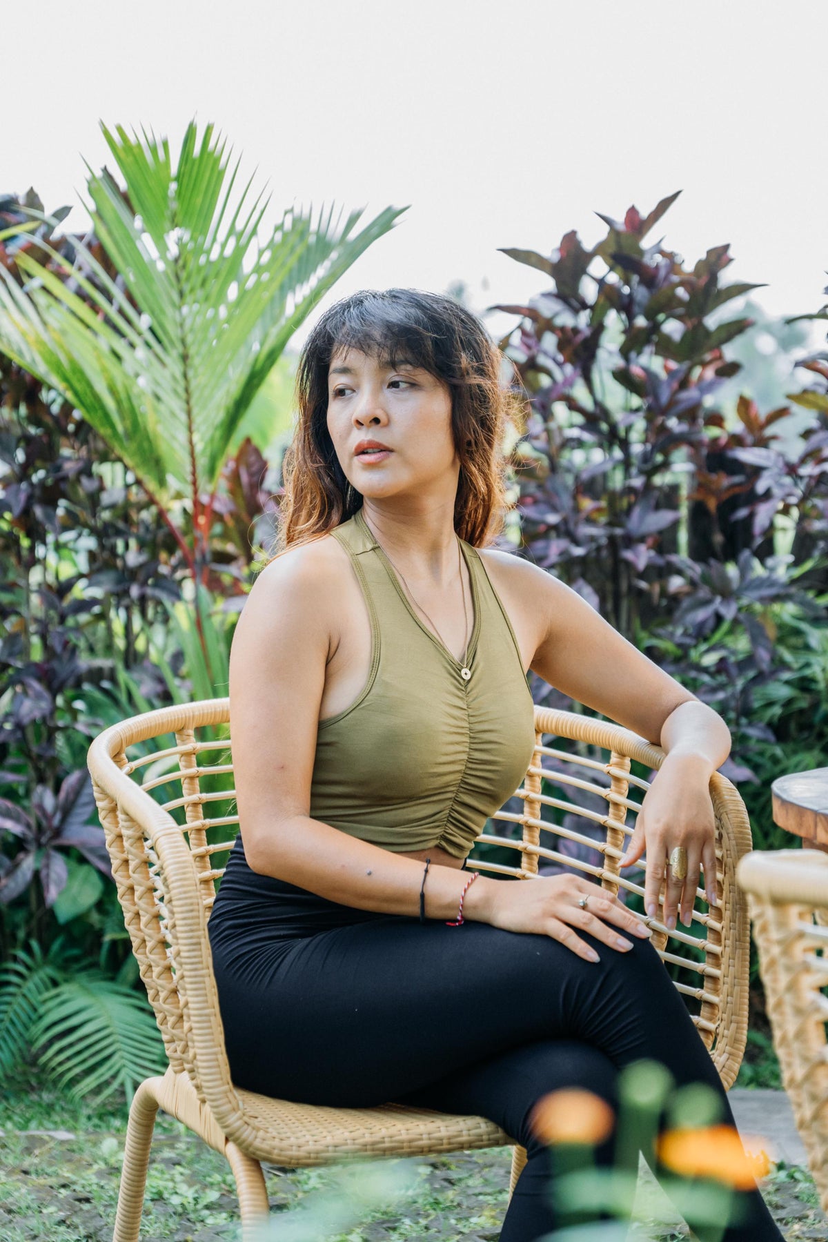 A woman wearing a sage green ruched tank top and black leggings sits in a woven rattan chair in an outdoor garden setting with lush green and purple foliage.