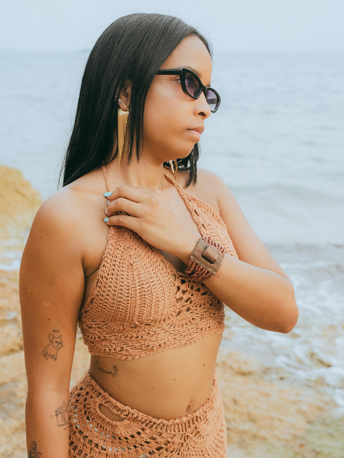 A woman wearing a matching crochet halter top and skirt set in a warm tan color, standing on a beach with the ocean in the background.