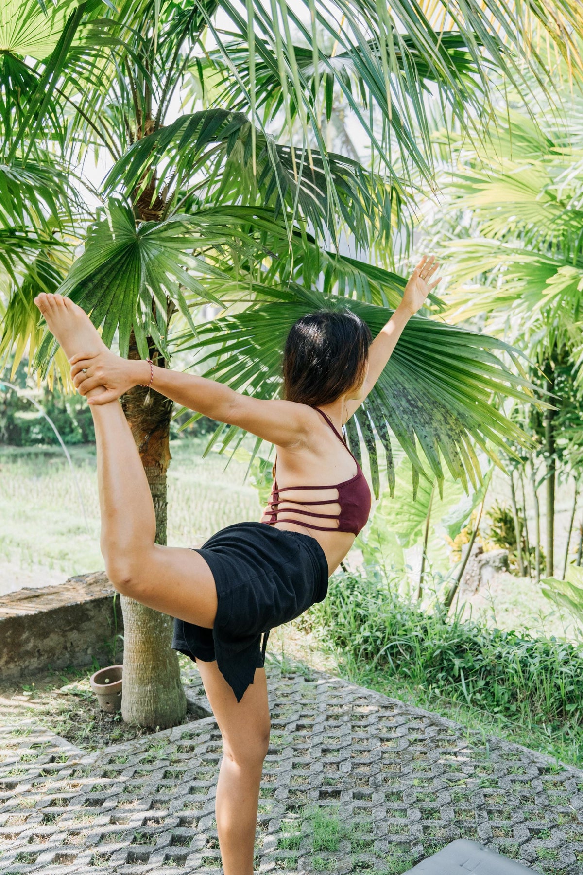 A woman in a maroon strappy sports bra and black shorts performs a yoga pose outdoors on a stone path surrounded by lush green palm trees.