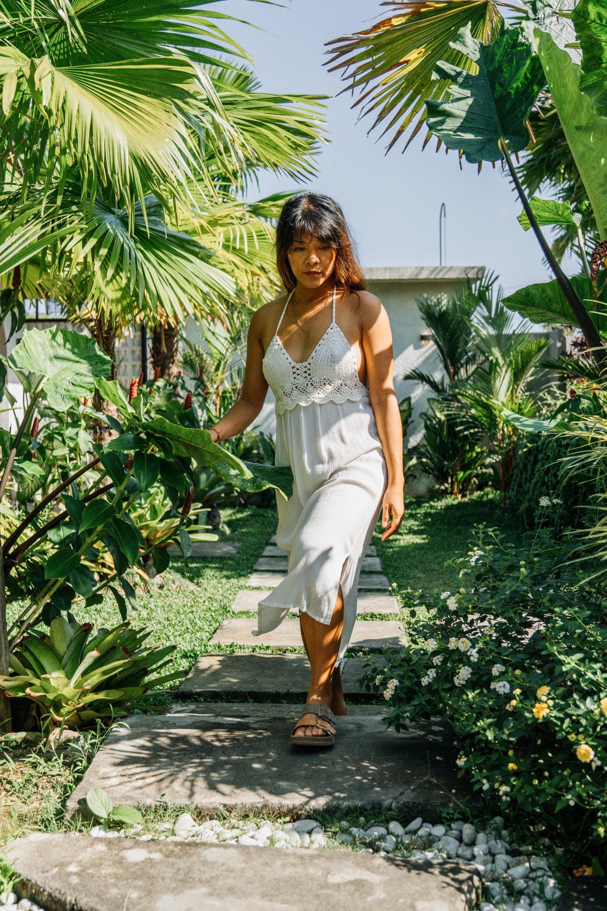 A woman wearing a white crochet halter dress walks down a stone path surrounded by lush tropical green plants and garden foliage.