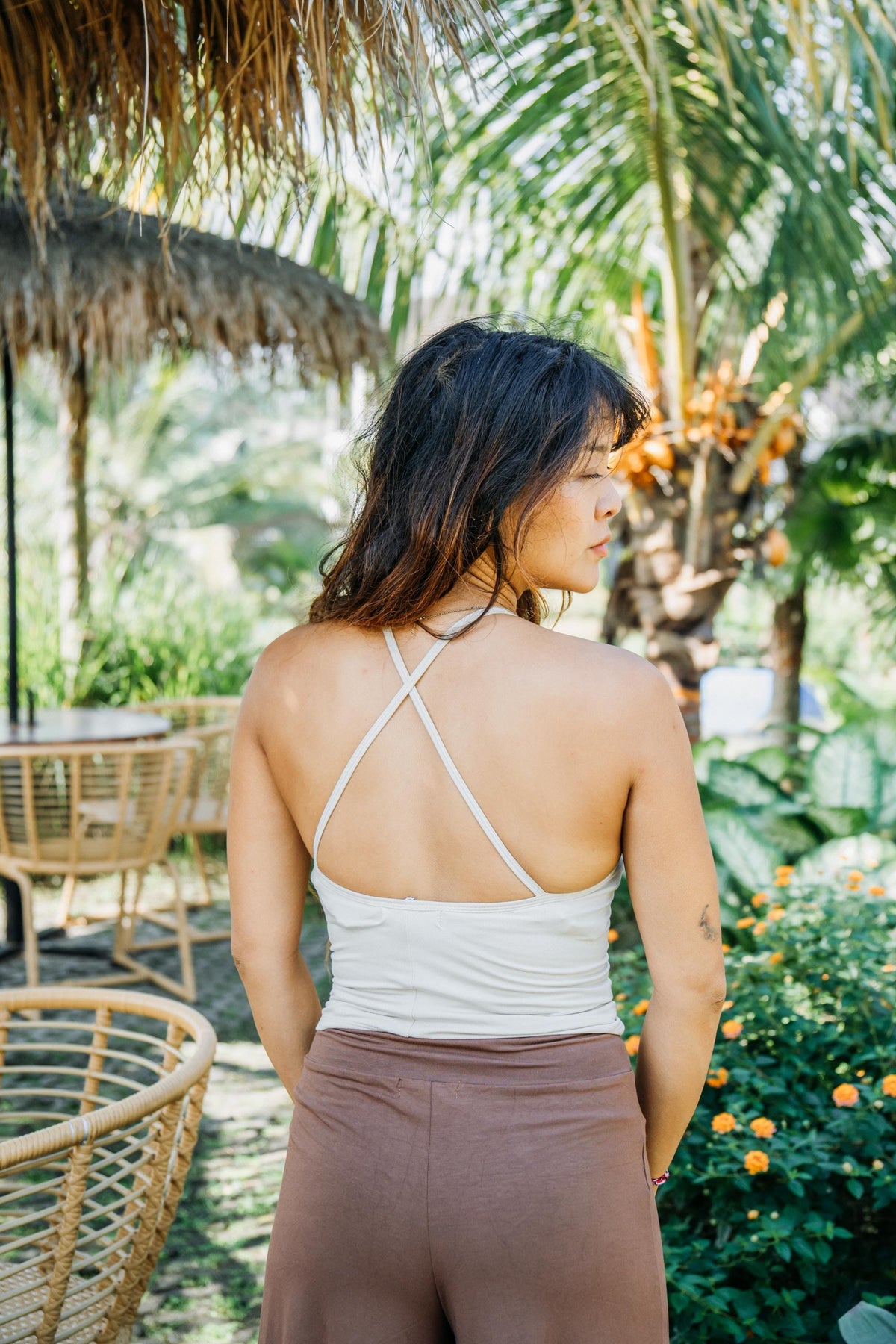 A woman is shown from behind wearing a white strappy halter-style tank top and brown leggings in an outdoor tropical garden setting with palm trees and a thatched umbrella.