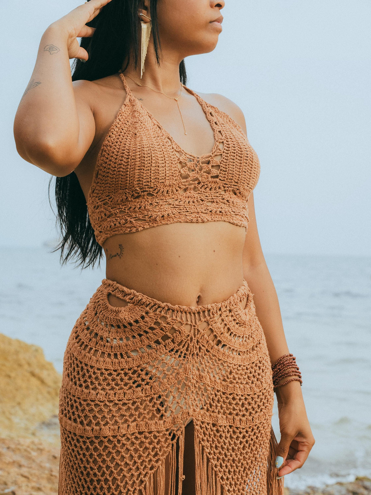 A woman wearing a matching crochet bikini-style top and long skirt in a warm rust tan color, standing outdoors near the ocean with rocks in the background.