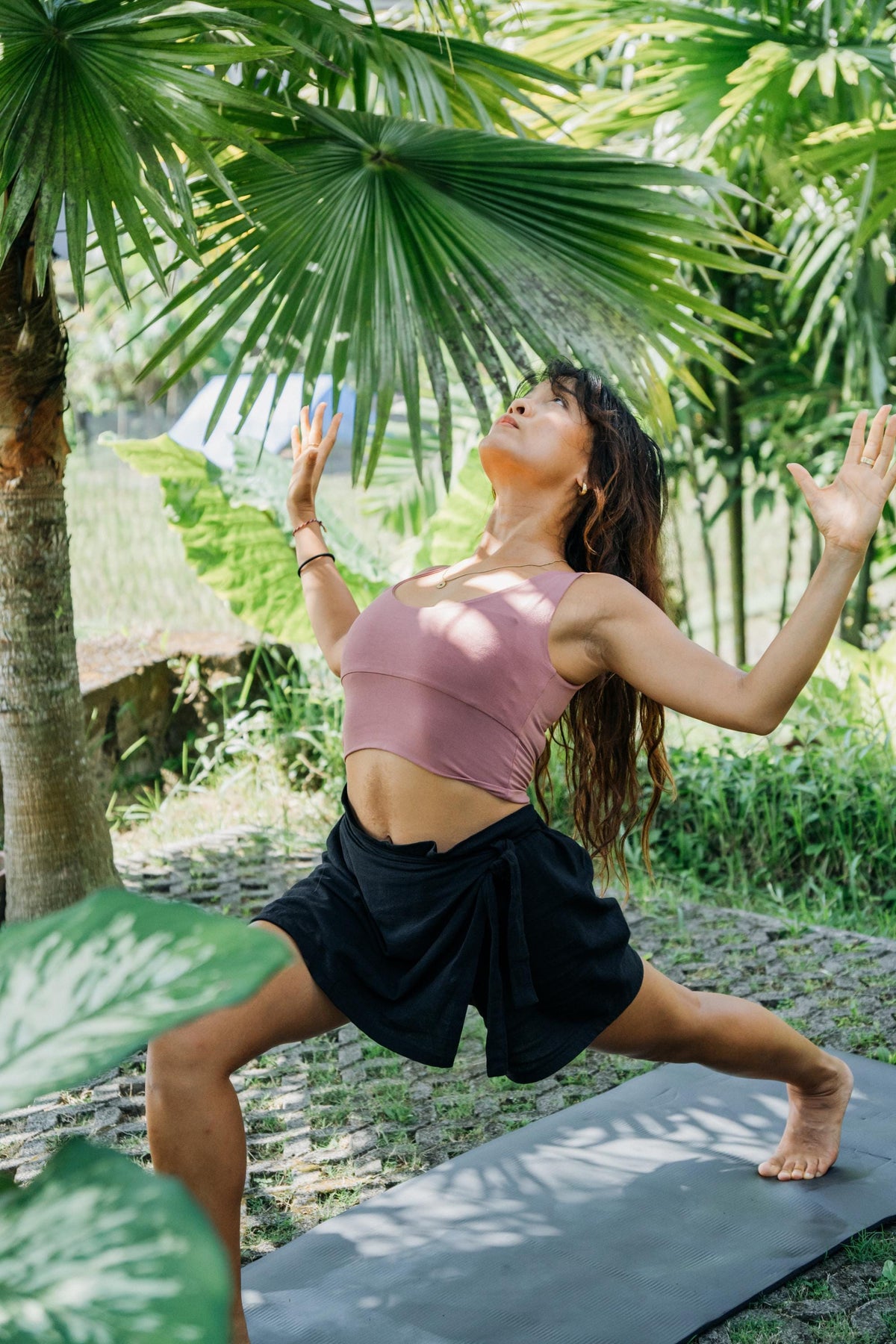 A woman in a pink sports bra and black wrap skirt does a yoga pose on a dark yoga mat outdoors among lush green palm trees.
