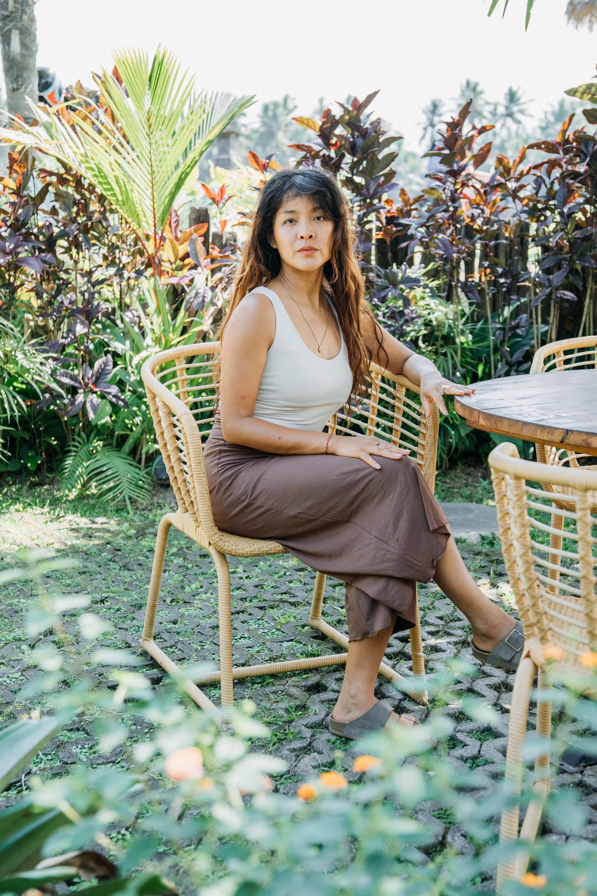A woman sits on a woven rattan chair in a lush garden patio setting, wearing a light beige tank top and a brown midi skirt.