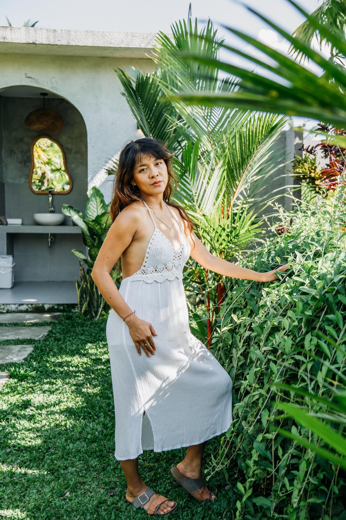 A woman wearing a white halter-neck maxi dress with crochet detailing stands in a lush garden setting next to a rustic building.