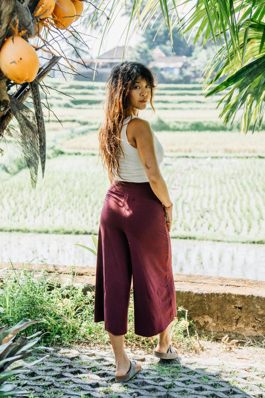A woman stands outdoors in front of green rice paddies, wearing a white sleeveless top and high-waisted, wide-leg burgundy cropped pants. She is looking back over her shoulder.