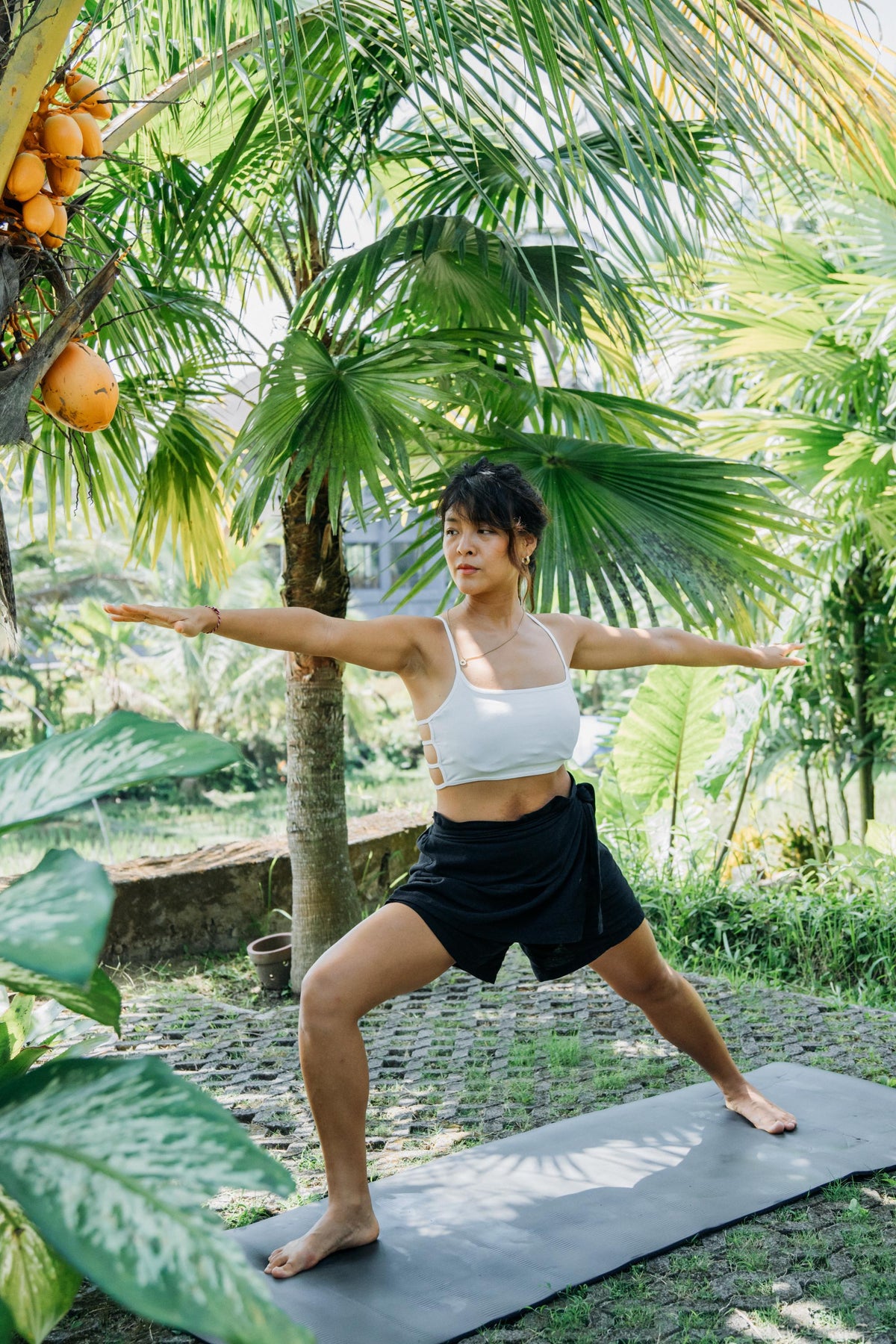 A woman in a white sports bra and black shorts performs a yoga pose on a gray mat in a lush tropical garden with palm trees and coconuts.