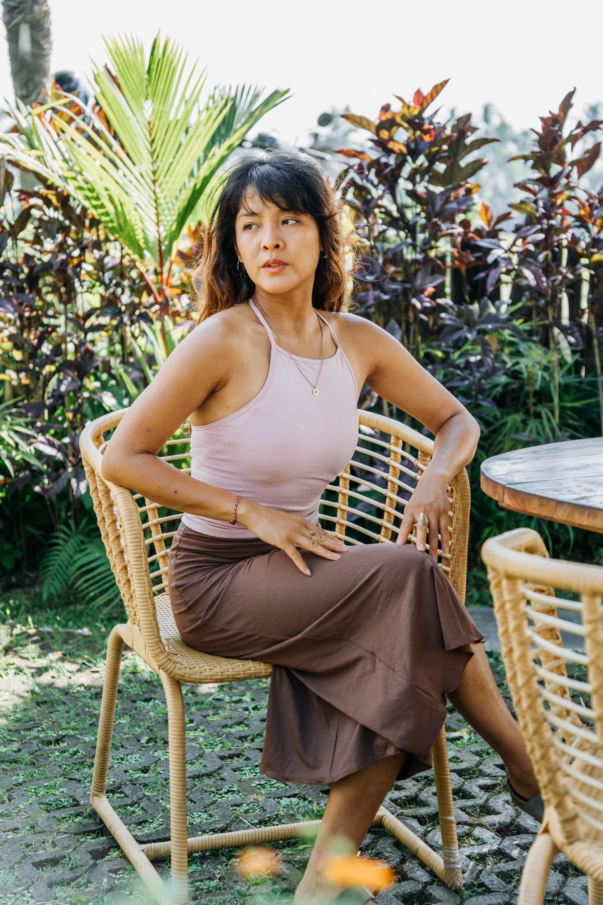 A woman sits on a woven rattan chair in an outdoor garden setting, wearing a light pink halter-neck tank top and a brown midi skirt.