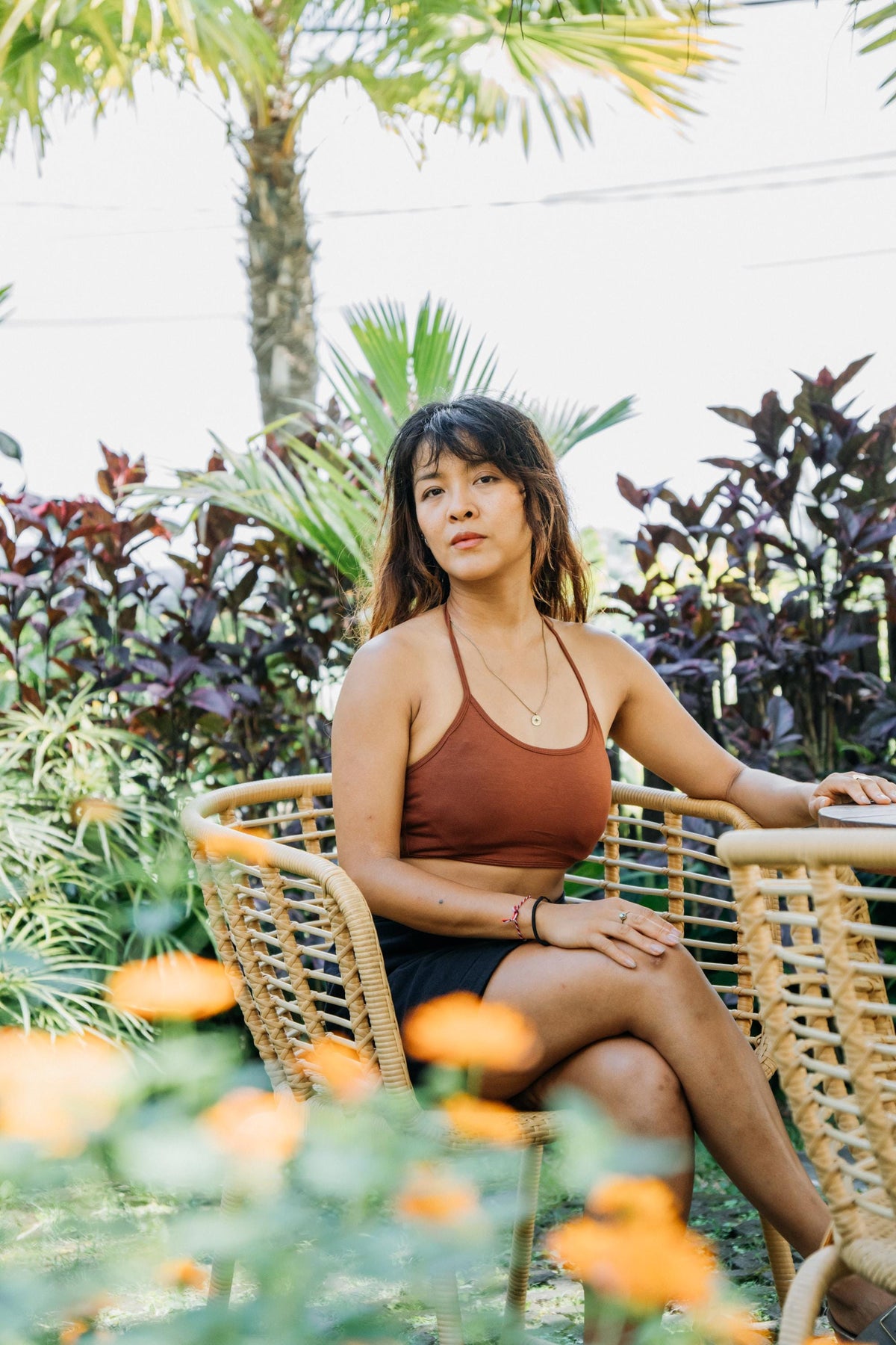 A woman sits in a wicker chair in a garden setting, wearing a rust-brown halter crop top and a black skirt.