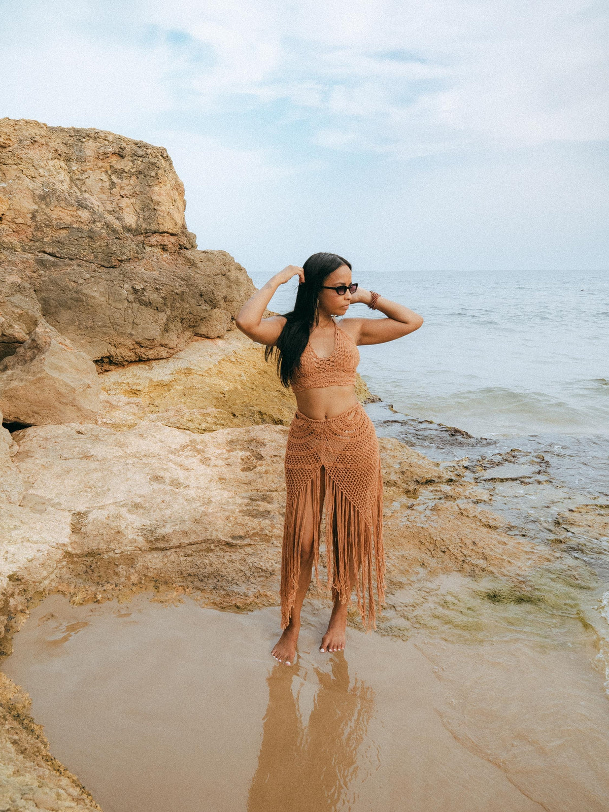 A woman stands barefoot in shallow water on a rocky beach, wearing a tan crochet halter bikini top and matching fringe sarong skirt.