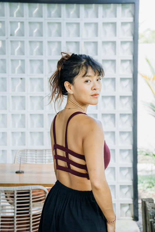 A woman wearing a dark burgundy strappy crop top with an open back and a black skirt, standing in front of a white glass block wall.