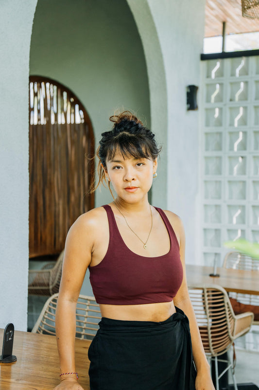 A woman with her hair in a bun stands in a cafe or restaurant setting, wearing a dark burgundy sports bra and a black skirt tied at the waist.