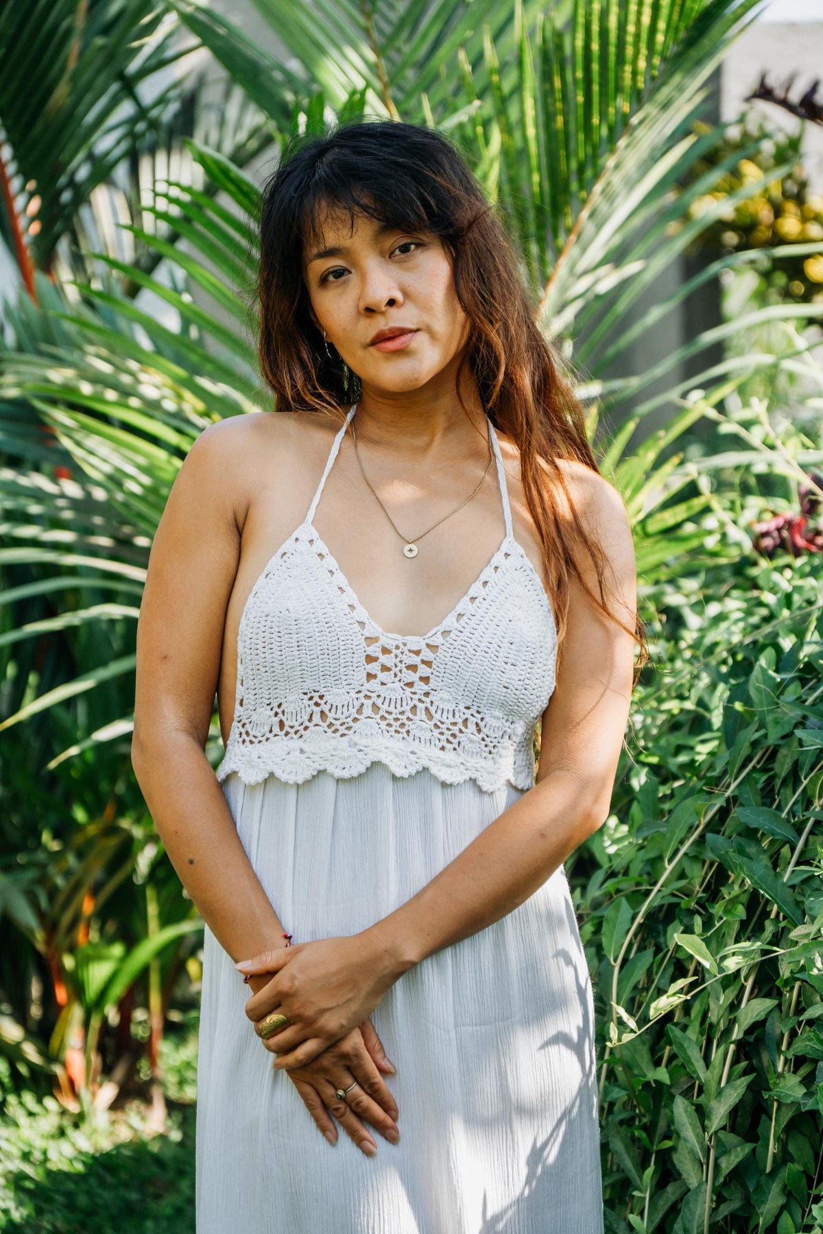 A woman wearing a white crochet halter top and matching white pleated skirt, standing outdoors in front of lush green tropical foliage.