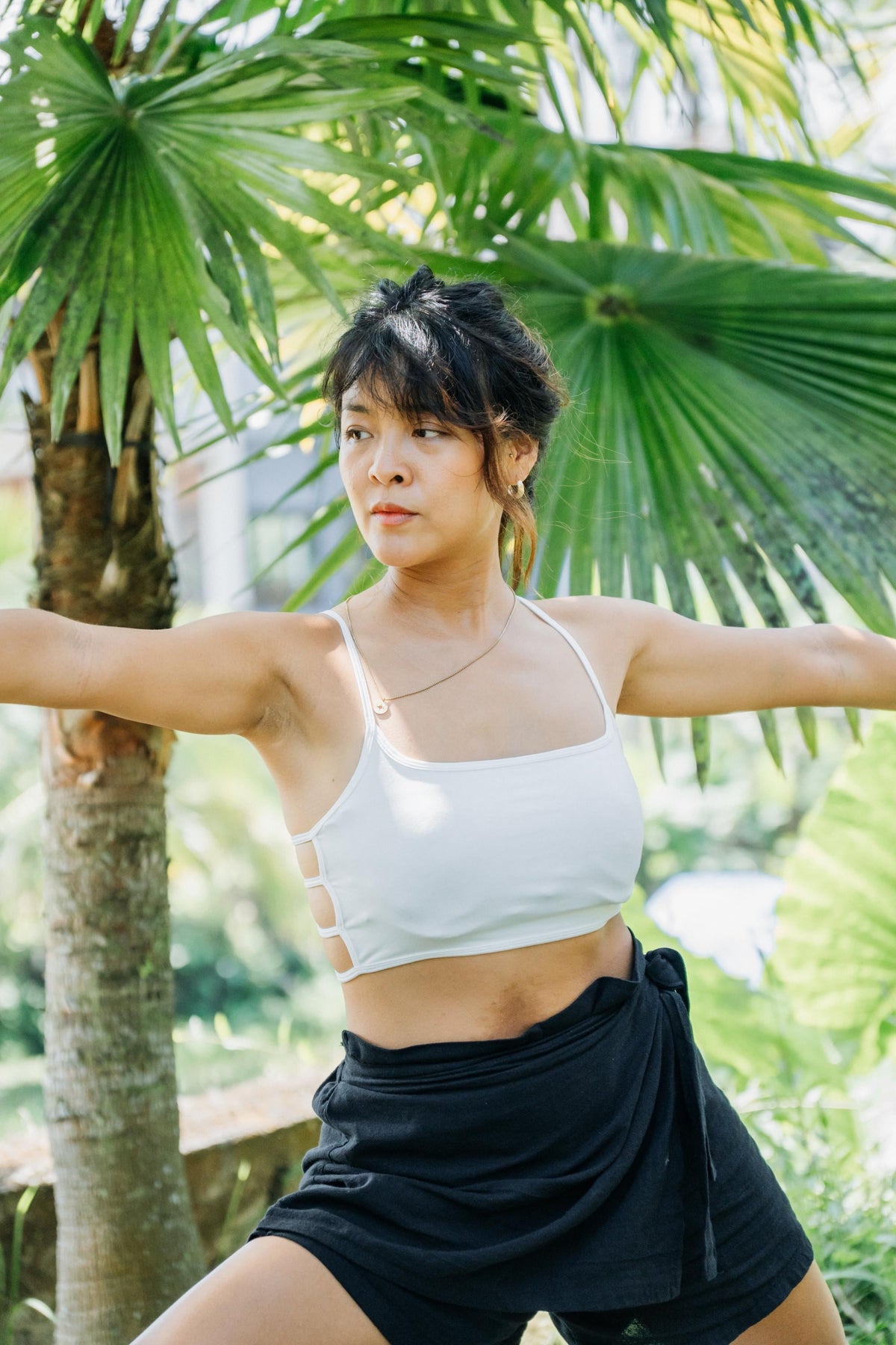 A woman outdoors wearing a white cropped tank top with side cutouts and black wrap-style shorts, posed with arms outstretched in front of large green palm leaves.