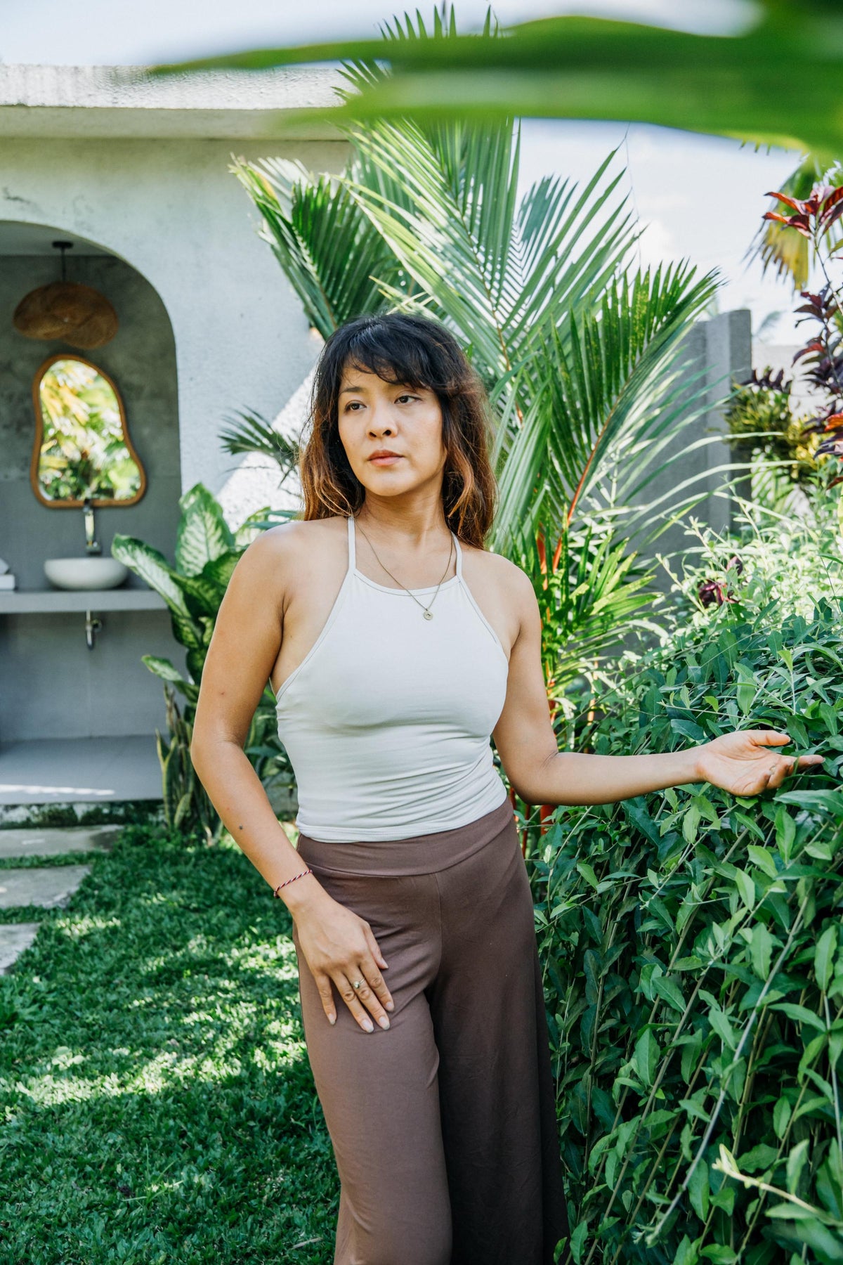 A woman stands outdoors in a garden setting wearing a white halter top and brown wide-leg pants, with lush green plants and a stone structure in the background.