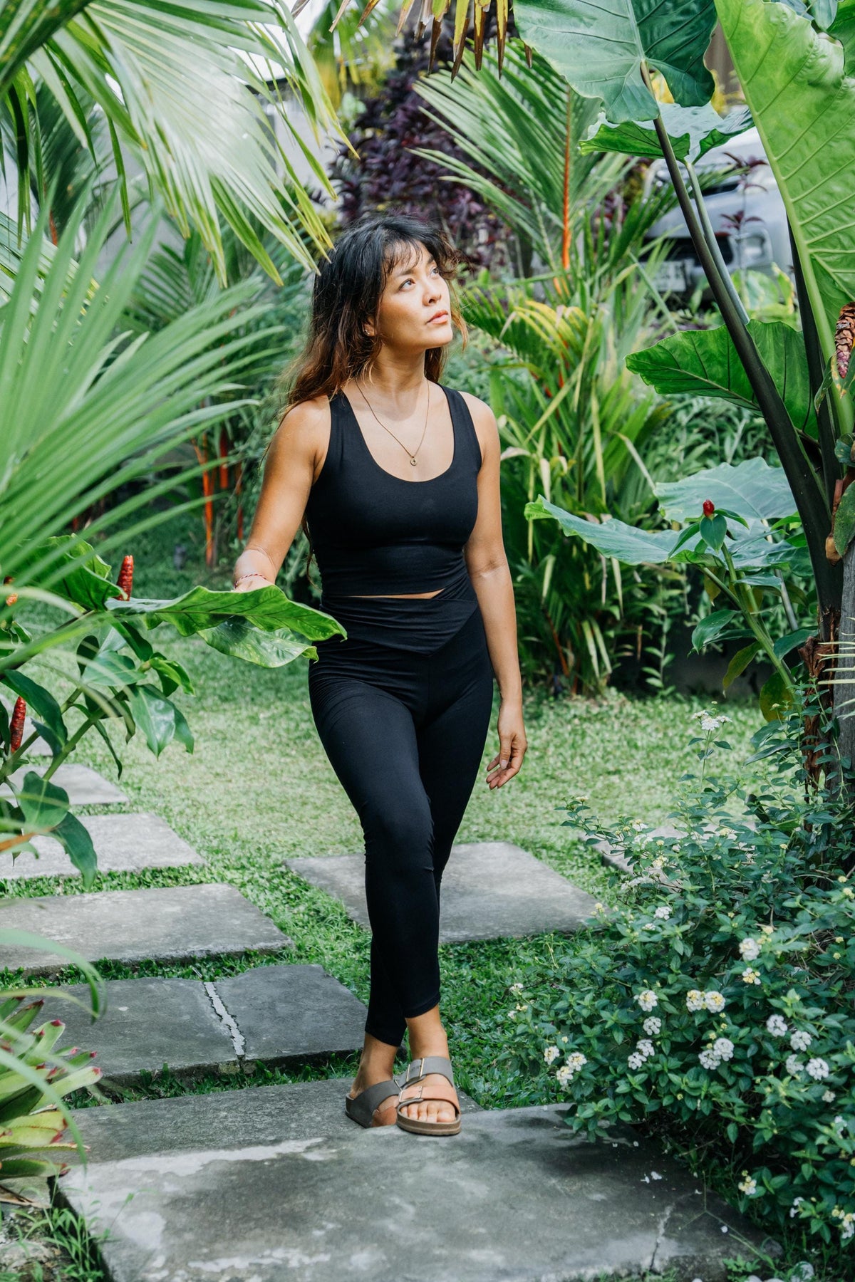 A woman wearing a black two-piece activewear set consisting of a tank top and leggings, walking along a stone path surrounded by lush tropical green plants.