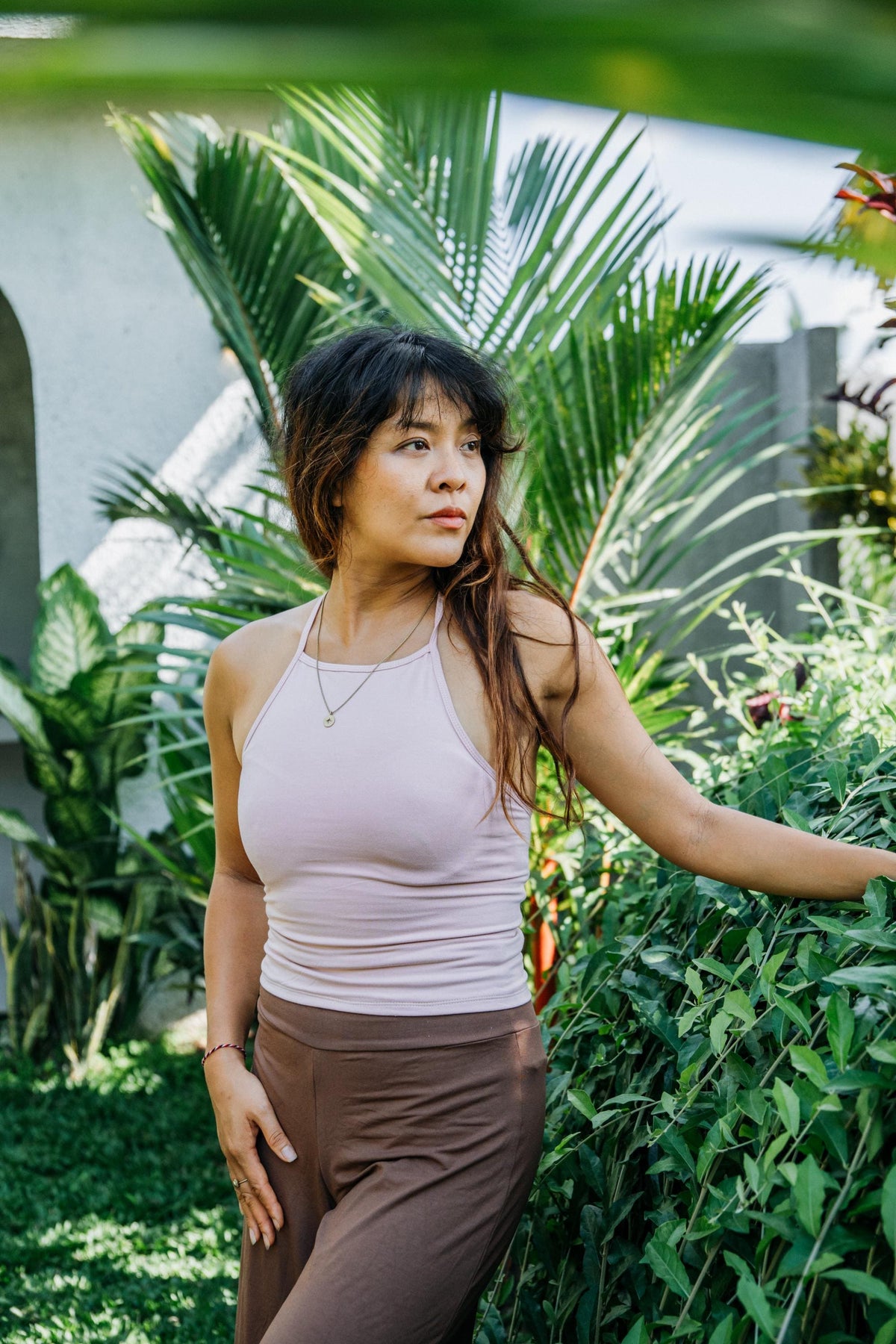 A woman posing outdoors in a garden setting, wearing a light pink ribbed halter tank top and brown pants.