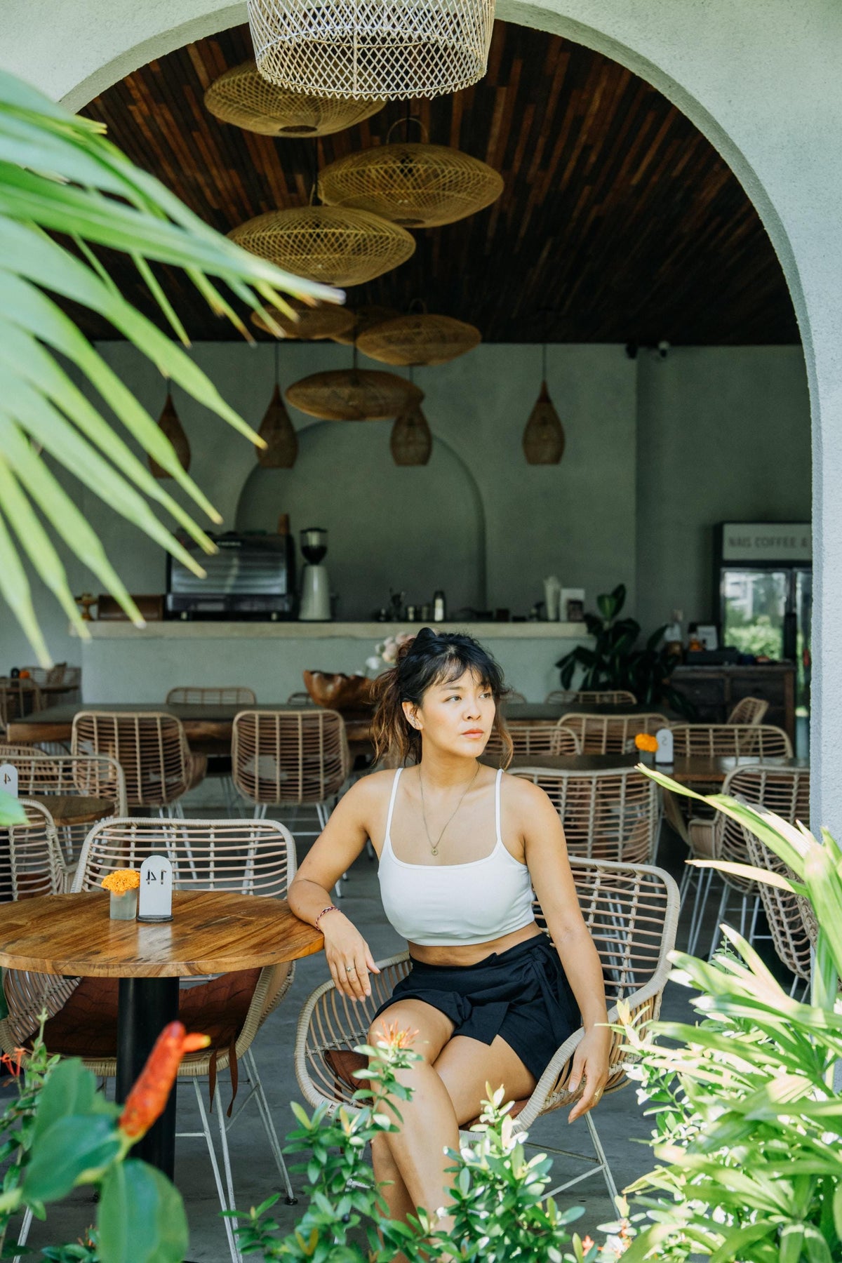 A woman sits at a round wooden table in a cafe, wearing a white ribbed tank top and black shorts. The setting features woven wicker chairs, hanging woven pendant lights, and lush green tropical plants framing the scene.