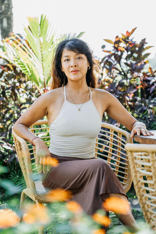 A woman sits in a woven rattan chair outdoors in a garden setting, wearing a beige halter top and a brown skirt.