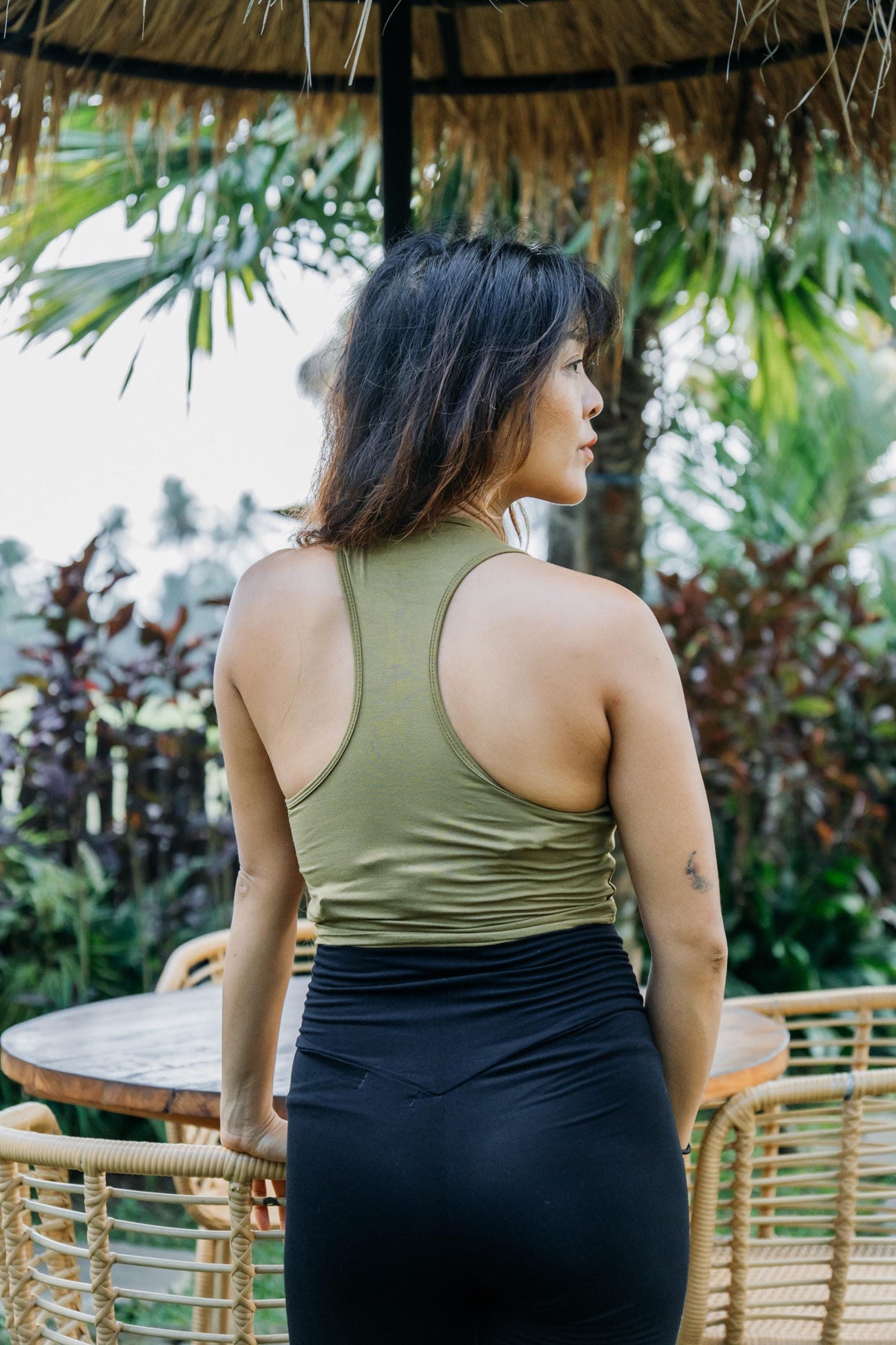 A woman is shown from behind wearing an olive green athletic tank top and a black high-waisted skirt, standing in a tropical patio setting with a thatched umbrella and wicker furniture.