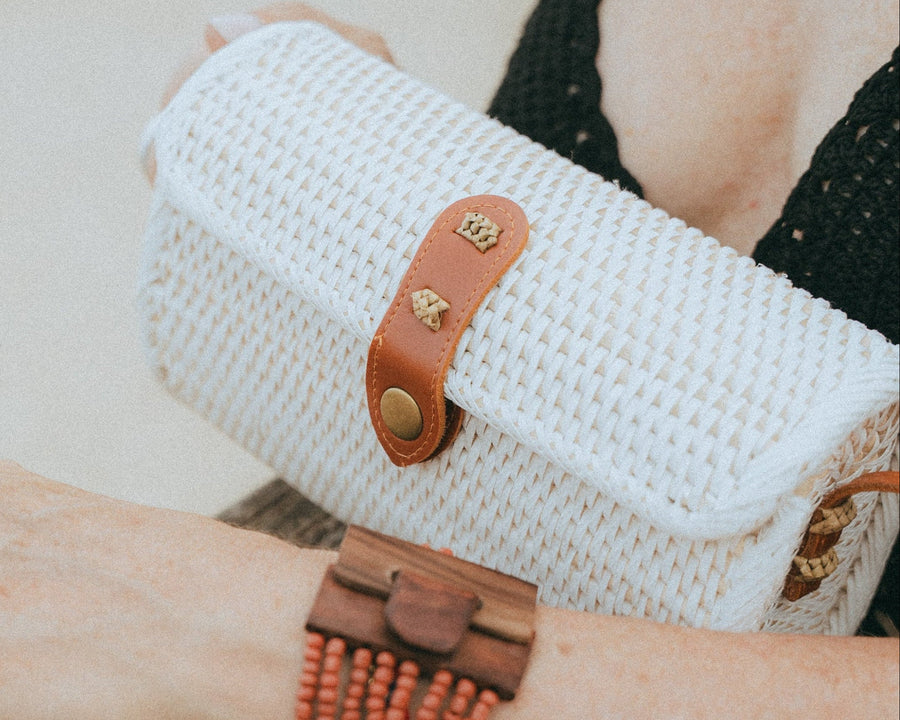 A close-up lifestyle shot of a person's arm wearing a brown wooden cuff bracelet with a beaded terracotta band, holding a white woven clutch bag with a brown leather strap.