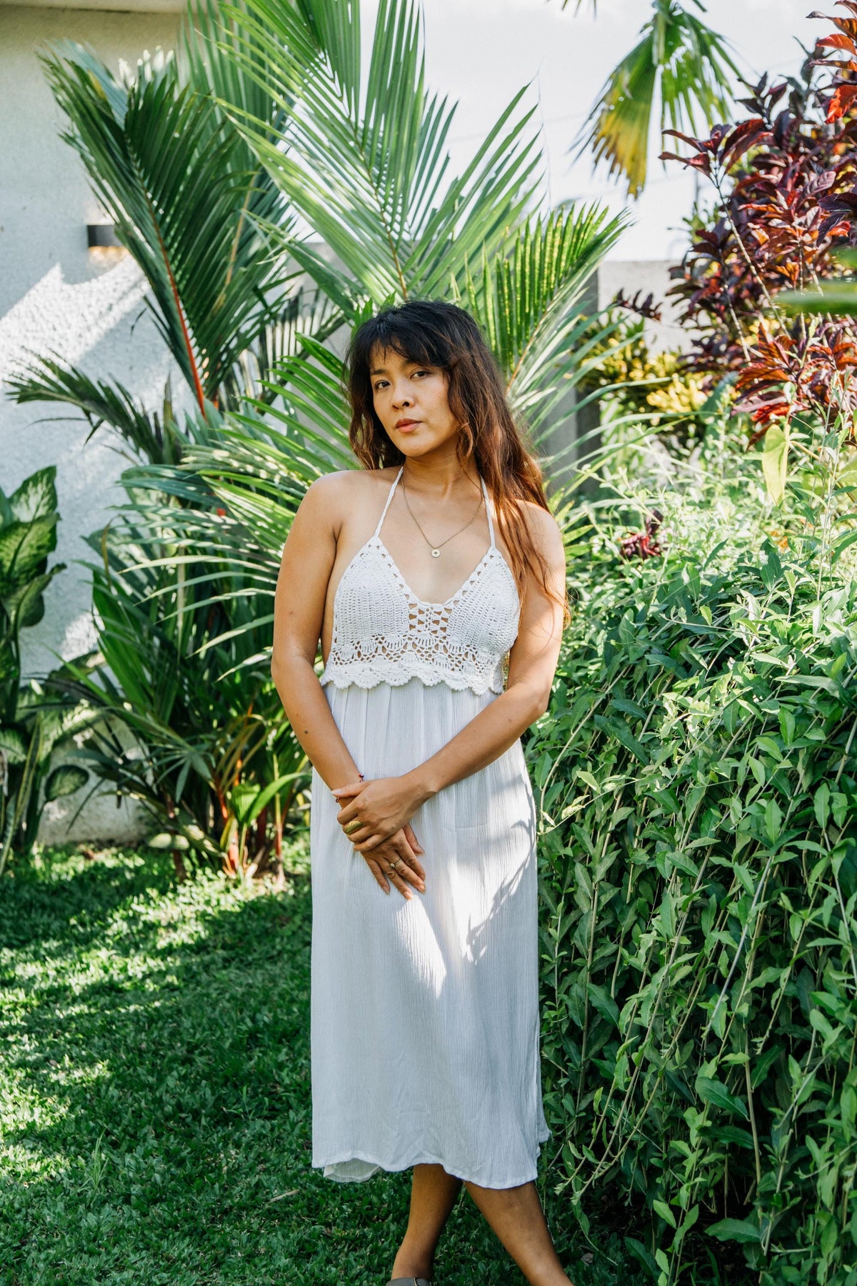 A woman wearing a white crochet halter midi dress stands outdoors in a lush garden with green plants and palm trees.