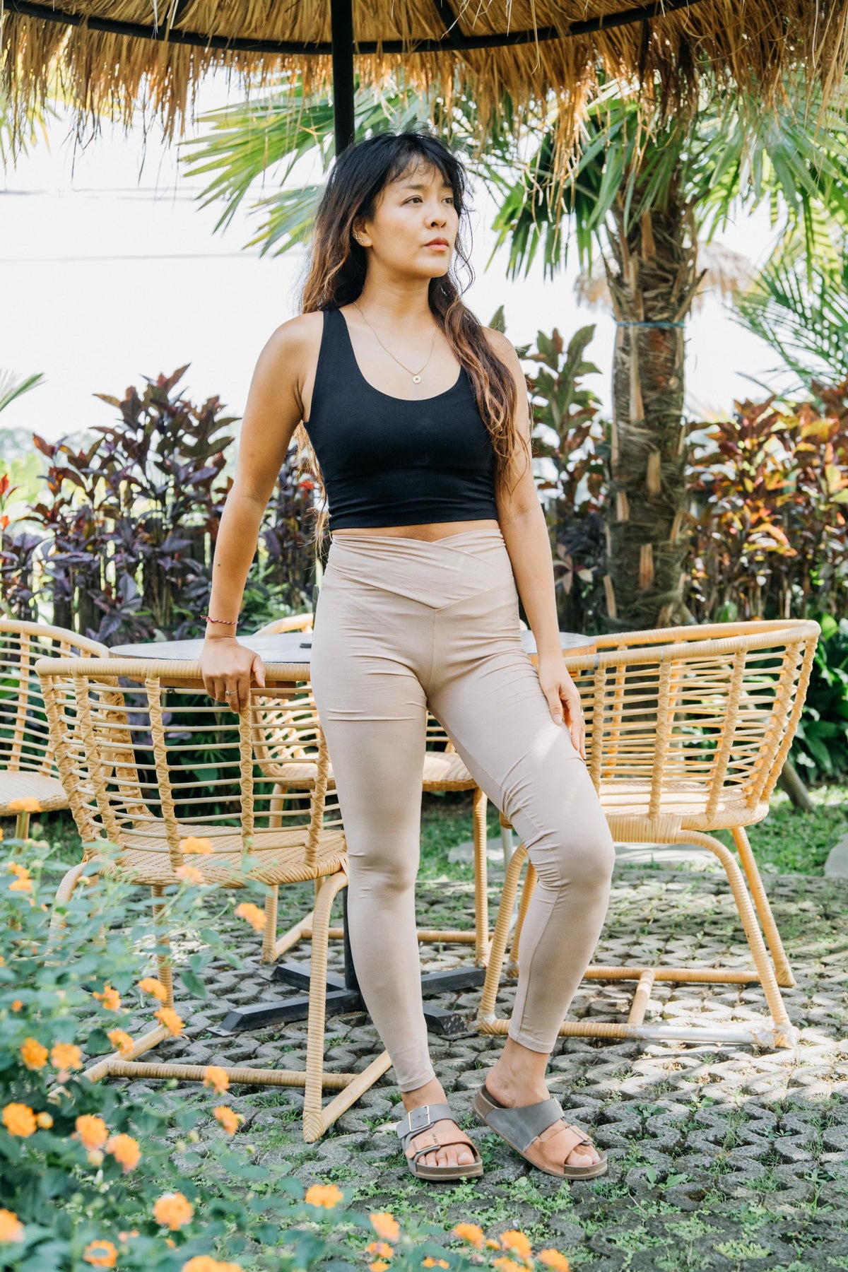 A woman stands outdoors in a garden patio setting wearing a black tank top and beige high-waisted leggings. She is leaning against a wicker chair under a thatched roof umbrella.