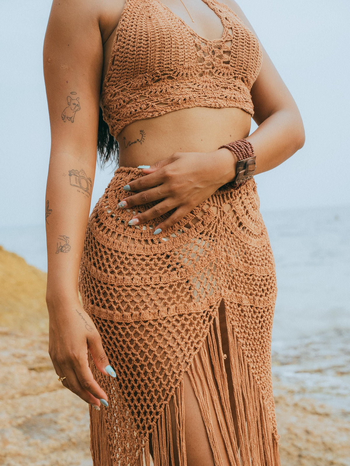 A woman wearing a matching crochet bikini top and high-waisted fringe skirt set in a warm terracotta or rust color, photographed outdoors near the beach with the ocean in the background.