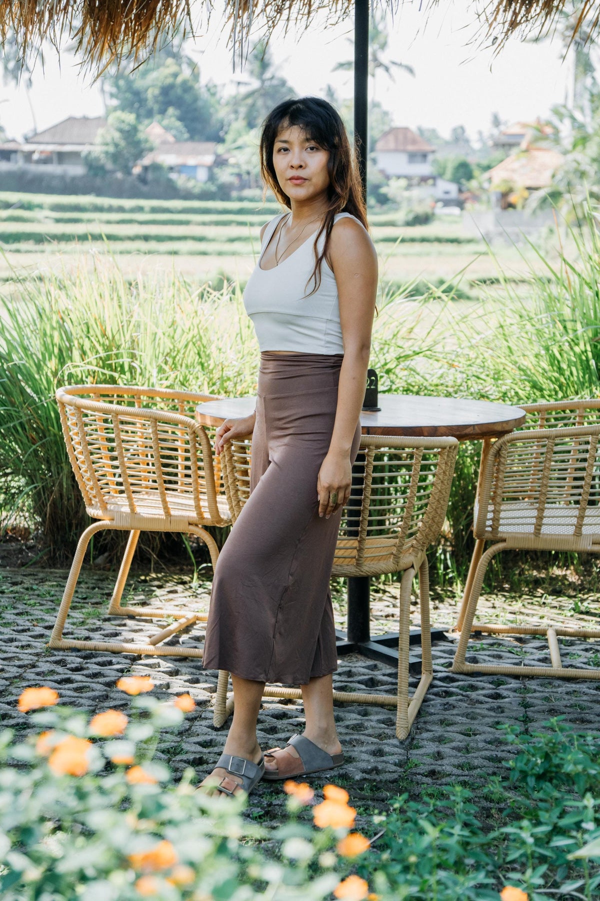 A woman stands outdoors in a tropical garden setting next to a wicker table and chairs, wearing a white sleeveless top and a brown midi skirt.