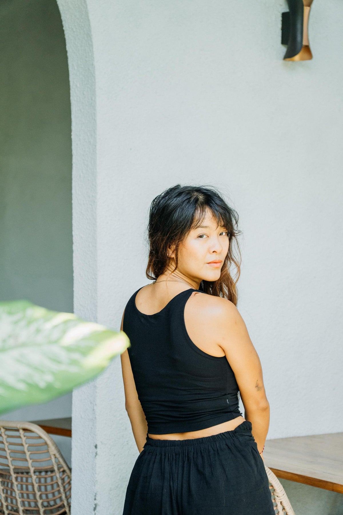 A woman with dark hair is shown from behind wearing a black sleeveless crop top and black skirt, standing in front of a light gray wall.