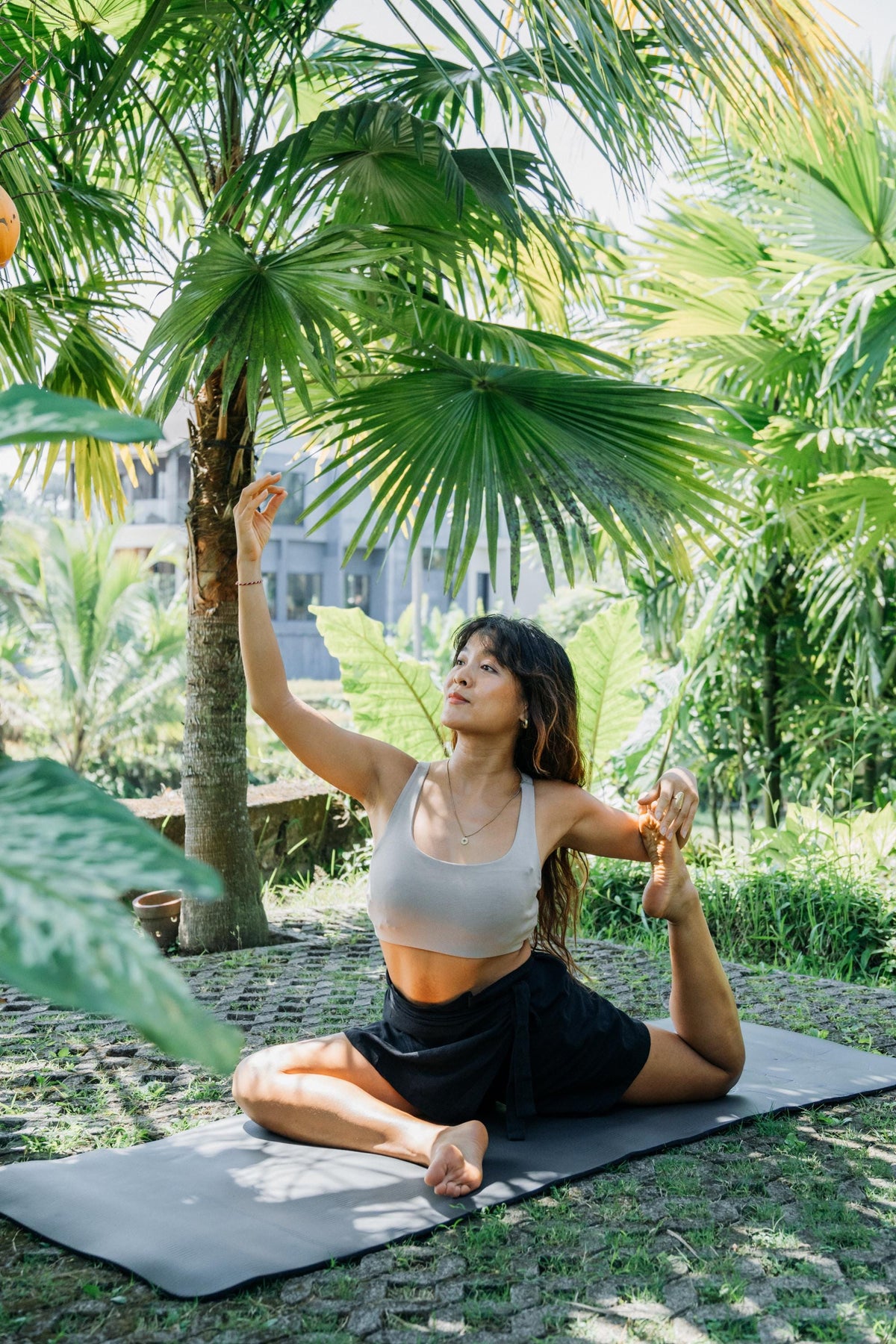 A woman in a light beige sports bra and black skirt performs a yoga pose on a gray mat in a lush tropical garden with palm trees.