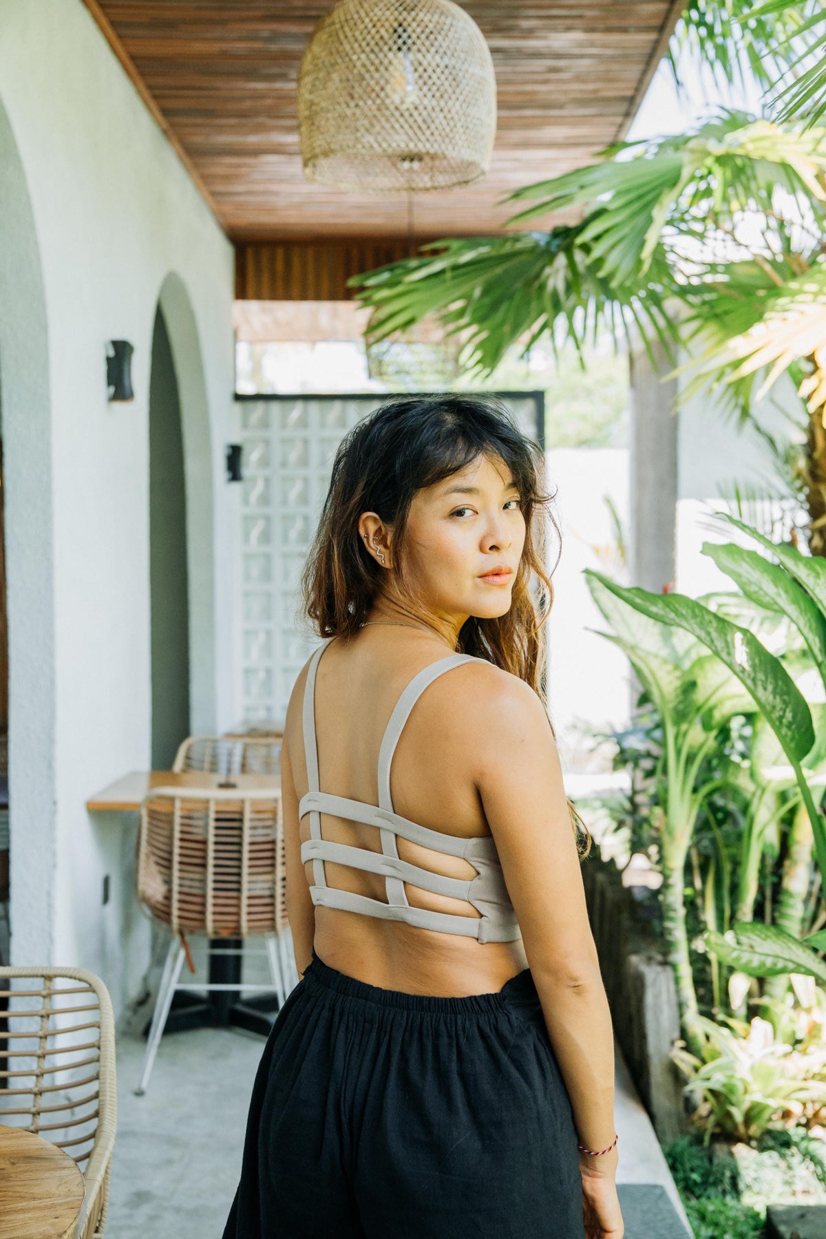 A woman wearing a light beige strappy cutout crop top and a black skirt, standing on a patio with plants and wicker furniture in the background.