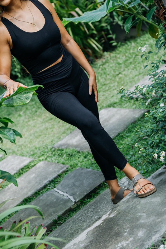 A woman wearing a black ribbed tank top and matching black leggings stands on outdoor stone steps surrounded by green plants and foliage.