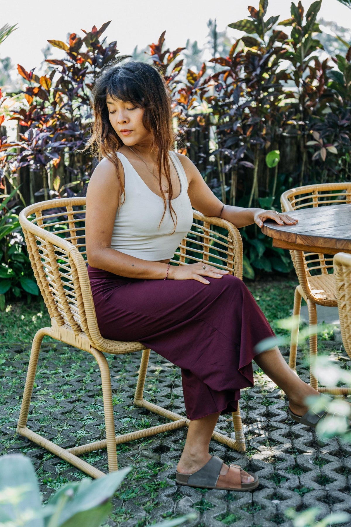 A woman sits on a woven rattan outdoor chair in a garden patio setting, wearing a white tank top, a long burgundy maxi skirt, and brown slide sandals.