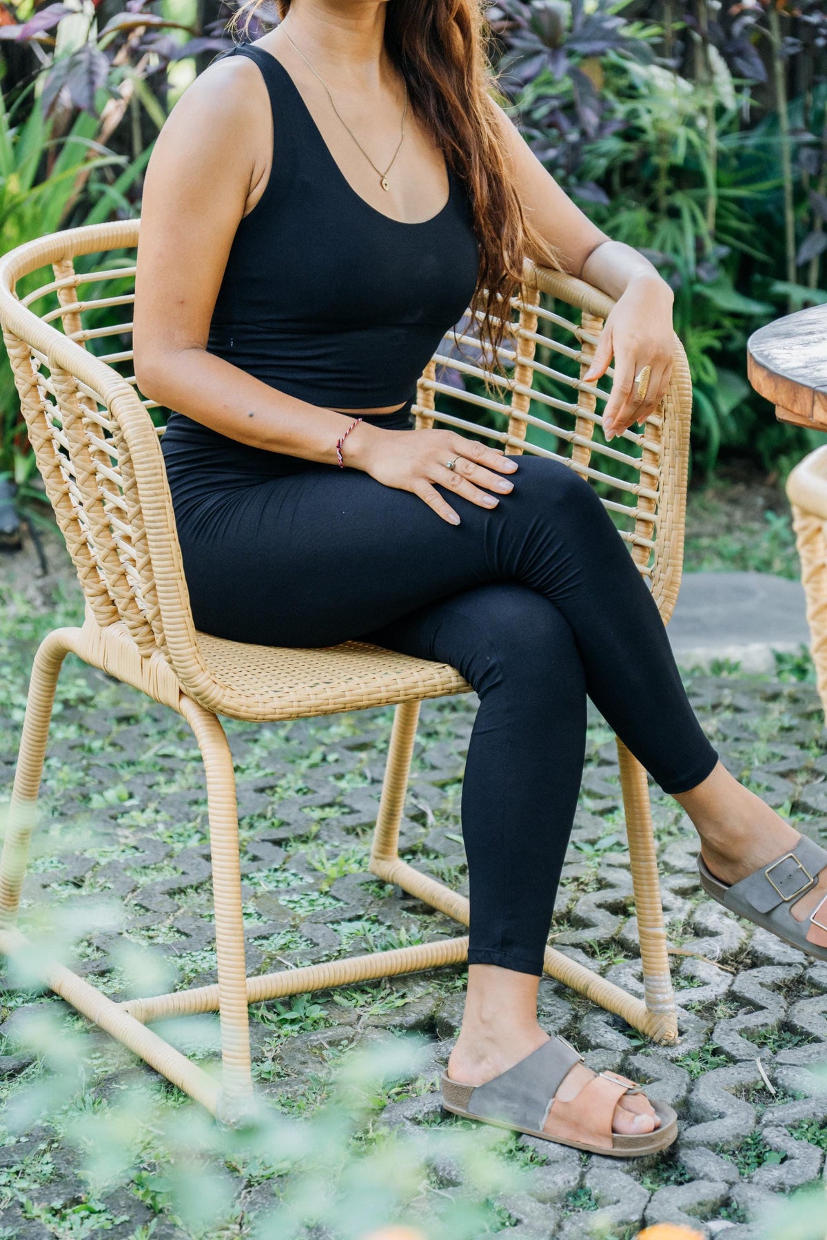 A woman wearing a black ribbed tank top and matching black leggings sits on a woven rattan outdoor chair in a garden setting.