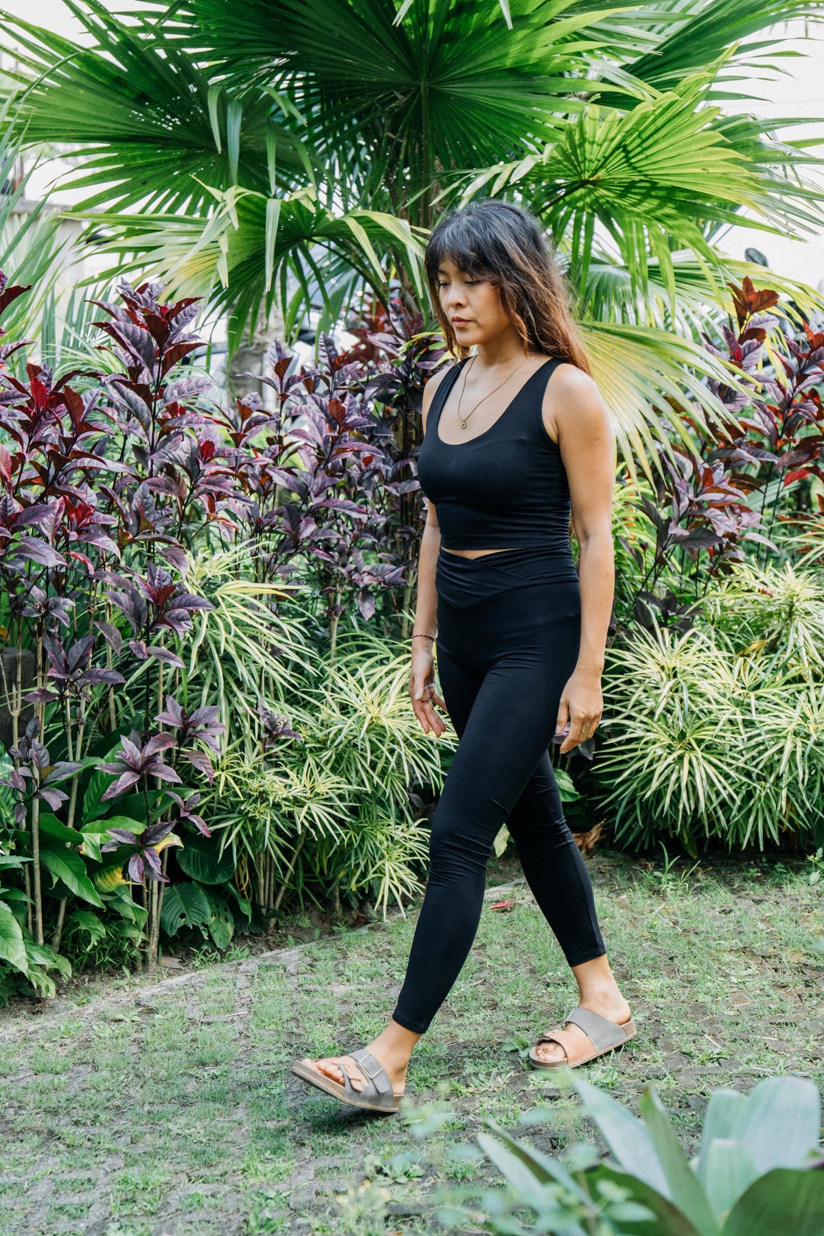 A woman wearing a matching black activewear set consisting of a scoop-neck tank top and high-waisted leggings, walking outdoors on a grassy path surrounded by lush tropical green and purple foliage.