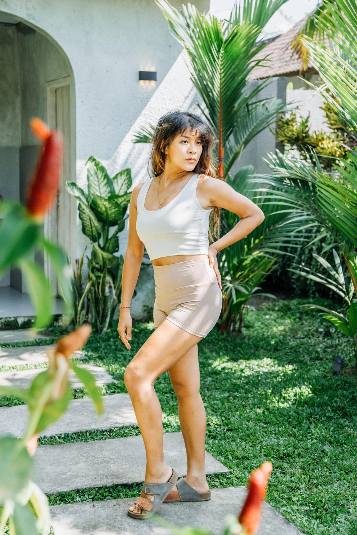 A woman posing outdoors in a tropical garden setting, wearing a matching beige two-piece activewear set consisting of a tank top and shorts.