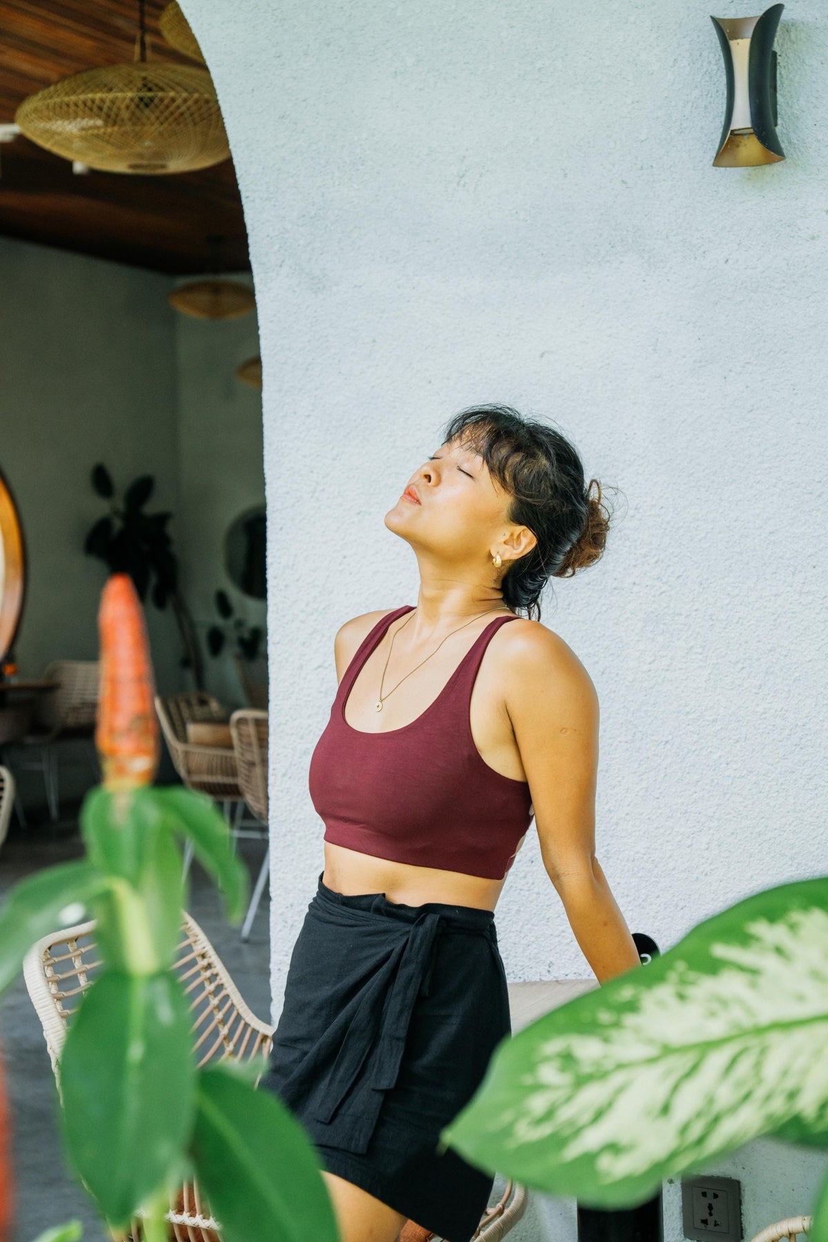A woman wearing a burgundy sports bra and black skirt ties her head back in surprise, standing in a sunlit patio setting with a textured light blue wall, plants, and wicker chairs in the background.